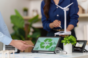 Team member points at a tablet showing ESG/recycling infographic amid a desk with a wind turbine model and green plant, symbolizing sustainability work.
