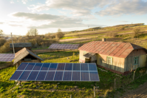 Sunlit rural farmyard with multiple solar panels on metal frames in front of wooden buildings and rolling hills in the distance.