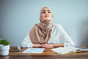 Professional woman wearing a tan hijab sits at a wooden desk with papers, a notebook, and glasses, looking confident against a light blue wall.