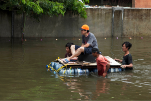 Four men on an improvised raft of plastic drums and wooden planks navigate brown floodwater; one wears an orange cap beside a concrete barrier.