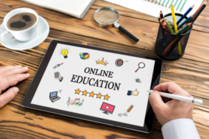 Tablet on a wooden desk shows a screen with 'Online Education' and icons (lightbulb, crown, stars, computer) as a person writes with a stylus.